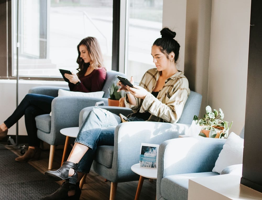 Two women using their gadgets while sitting on chairs